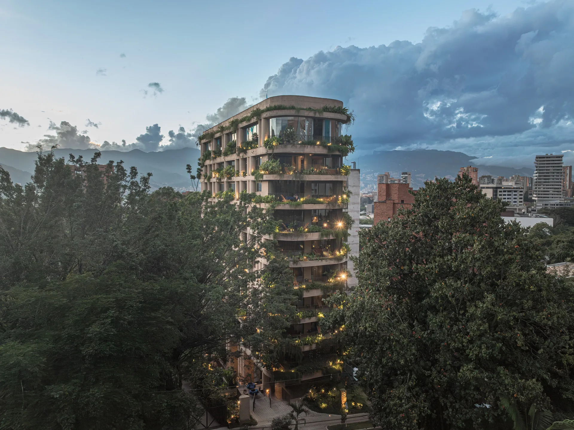 23 Living entre el arbolado de Medellín, con las montañas y un cielo de atardecer nublado al fondo.